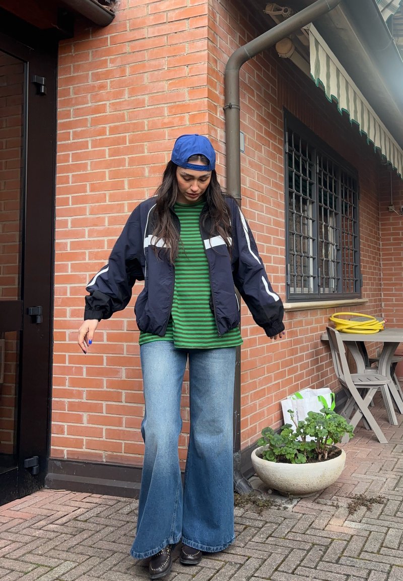 Frau trägt blaue Kappe rückwärts, marineblaue Jacke, grünes gestreiftes Hemd und weite Jeans, steht auf einem Ziegel-Terrasse nahe einem Topfpflanze und Gartenmöbeln.