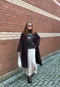 Femme portant un long manteau sombre, un pull camouflage, une jupe crème et des bottes, tenant un sac blanc, marchant sur une rue pavée à côté d'un mur en briques.