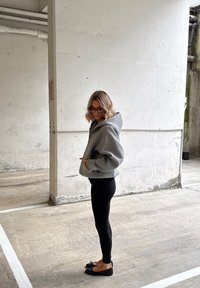 Kobieta o blond włosach do ramion, nosząca czarne okulary, szarą bluzę z kapturem, czarne legginsy i baleriny, stojąca w betonowym garażu podziemnym.