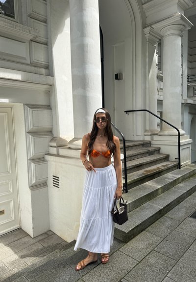 Top de bikini naranja, falda maxi blanca de volantes, gafas de sol transparentes, tela texturizada y un pequeño bolso marrón de mimbre, de pie en unos escalones de piedra.