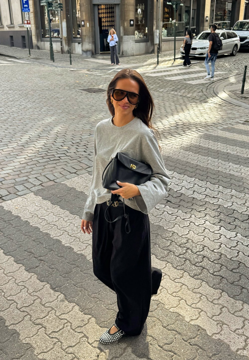 Sweatshirt cinza de manga longa, calças largas pretas, sapatos planos pretos com tachas, a carregar uma mala preta com detalhes em dourado, numa rua de calçada.