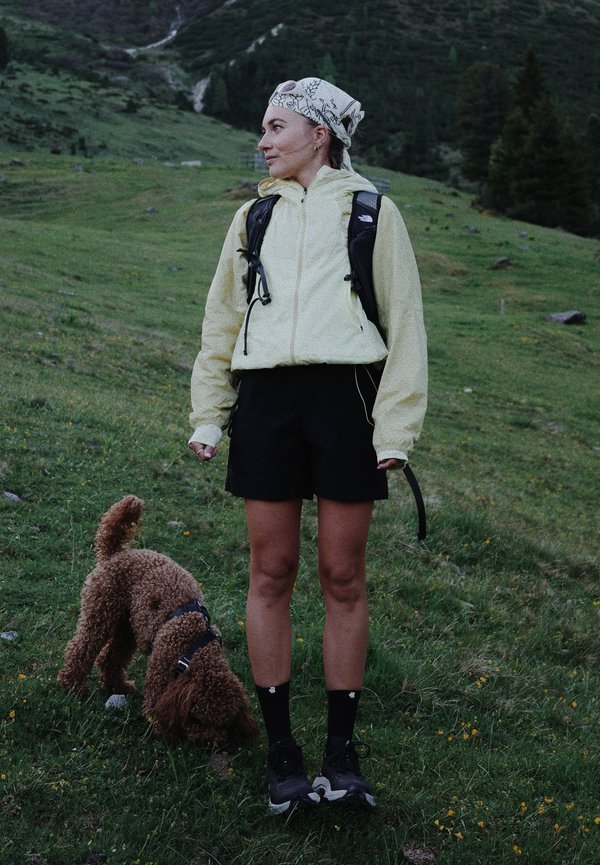 Femme en veste légère et short avec un foulard sur la tête, debout sur une colline herbeuse en randonnée avec un chien brun aux poils bouclés qui renifle le sol à côté d'elle.