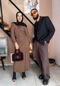 Καφέ ριμπ φόρεμα με μακριά μανίκια και ζώνη που δένει στη μέση; μαύρες διαφανείς μποτές με τακούνι; ψεύτικη γούνα τσάντα σε σκούρο καφέ χρώμα; άνδρας με μαύρη σακάκι και σκούρα παντελόνια.