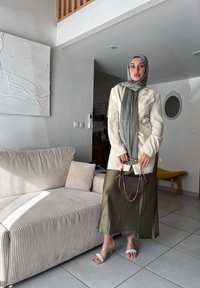 Κρεμ σακάκι, ελαιόχρωμη σατέν φούστα, διαφανή τακούνια με πέρλες, κρατώντας μια καφέ τσάντα με αλυσίδα. Ριγέ μπεζ καναπές στο φόντο.