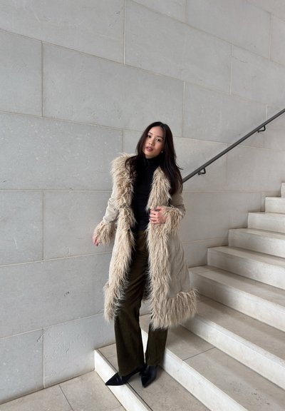 Mujer con un abrigo largo beige con ribete de piel, top negro y pantalones oscuros, de pie en unas escaleras de piedra clara junto a una pared con pasamanos de metal.