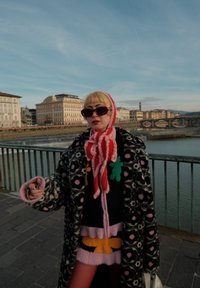 Persona con capelli biondi corti e frangia che indossa occhiali da sole scuri, cappotto a fantasia, sciarpa rossa e rosa, in piedi vicino a un fiume con lo sfondo di un paesaggio urbano.