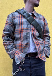 Veste à carreaux en gris, orange et marine avec une fermeture éclair. Bretelle matelassée verte traversant l'épaule. Porte-clés remarquable avec accessoires. Jean en denim.