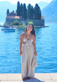 Beiger trägerloser Jumpsuit mit einer Schnüroptik an der Vorderseite, kombiniert mit einer handgefertigten Muschel-Handtasche. Der Hintergrund zeigt einen blauen See und Bäume.