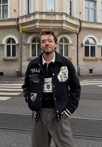 Veste varsity noire avec graphismes Yale, présentant des accents blancs, portée sur un t-shirt noir avec un logo. Un pantalon gris complète la tenue.