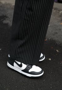 Černobílé tenisky Nike mají hladký design s děrovanou bílou špičkou, černými akcenty a strukturovanou podrážkou; nošeny s kalhotami s pruhy.