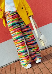 Kleurige wijde broek met strepen in levendige tinten roze, groen, blauw en zwart, gecombineerd met een gele jas en witte sneakers.