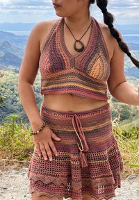 Femme portant un haut style crochet multicolore à dos nu et une jupe assortie, debout en plein air avec des montagnes et de la végétation en arrière-plan.
