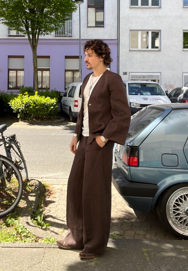 Man met krullend haar, gekleed in een bruin wijd broekpak en wit T-shirt, staat op de stoep naast geparkeerde auto's en fietsen.