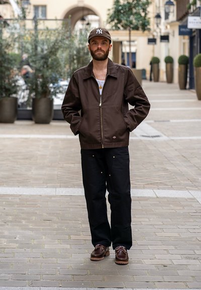 Chaqueta marrón de cremallera con cuello, combinada con pantalones anchos negros y zapatos marrones con cordones. Los accesorios incluyen una gorra y collares en capas.
