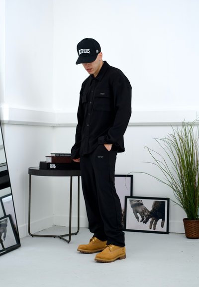 Camisa de manga larga negra y pantalones con la marca "TPDY", combinados con botas de trabajo color caqui, sobre un fondo minimalista y una mesa espejada.