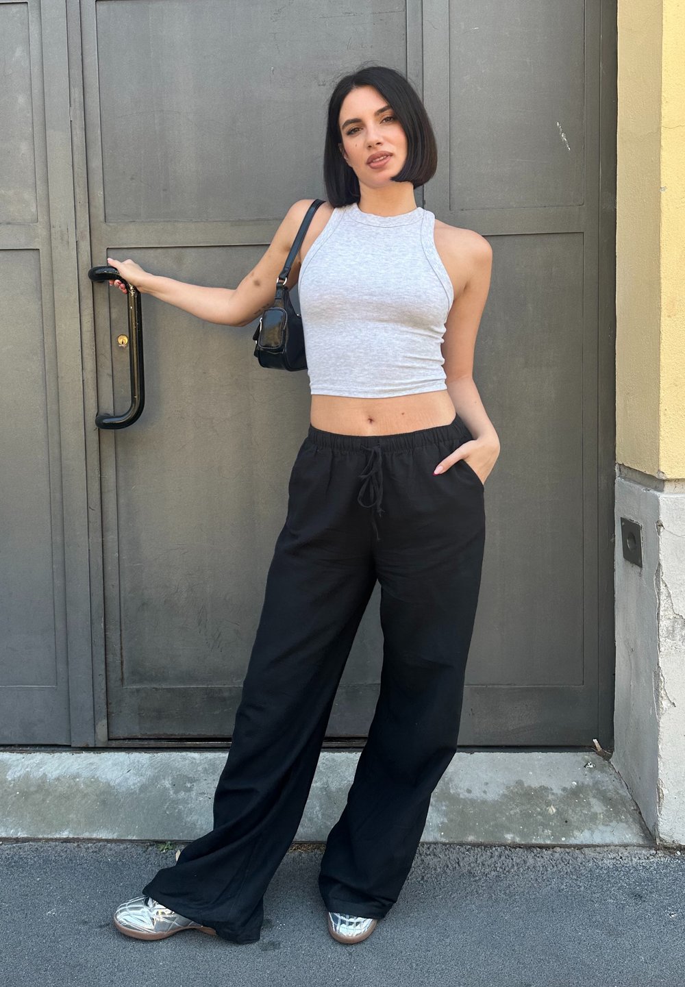 Šedý bezrukávový crop top, černé široké kalhoty s kapsami, stříbrné boty a malá černá kabelka, stojící u šedých dveří.