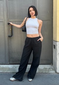 Šedý bezrukávový crop top, černé široké kalhoty s kapsami, stříbrné boty a malá černá kabelka, stojící u šedých dveří.