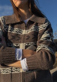 Femme portant un pull en maille marron et blanc à motifs, les bras croisés, debout à l'extérieur sous un ciel bleu clair.