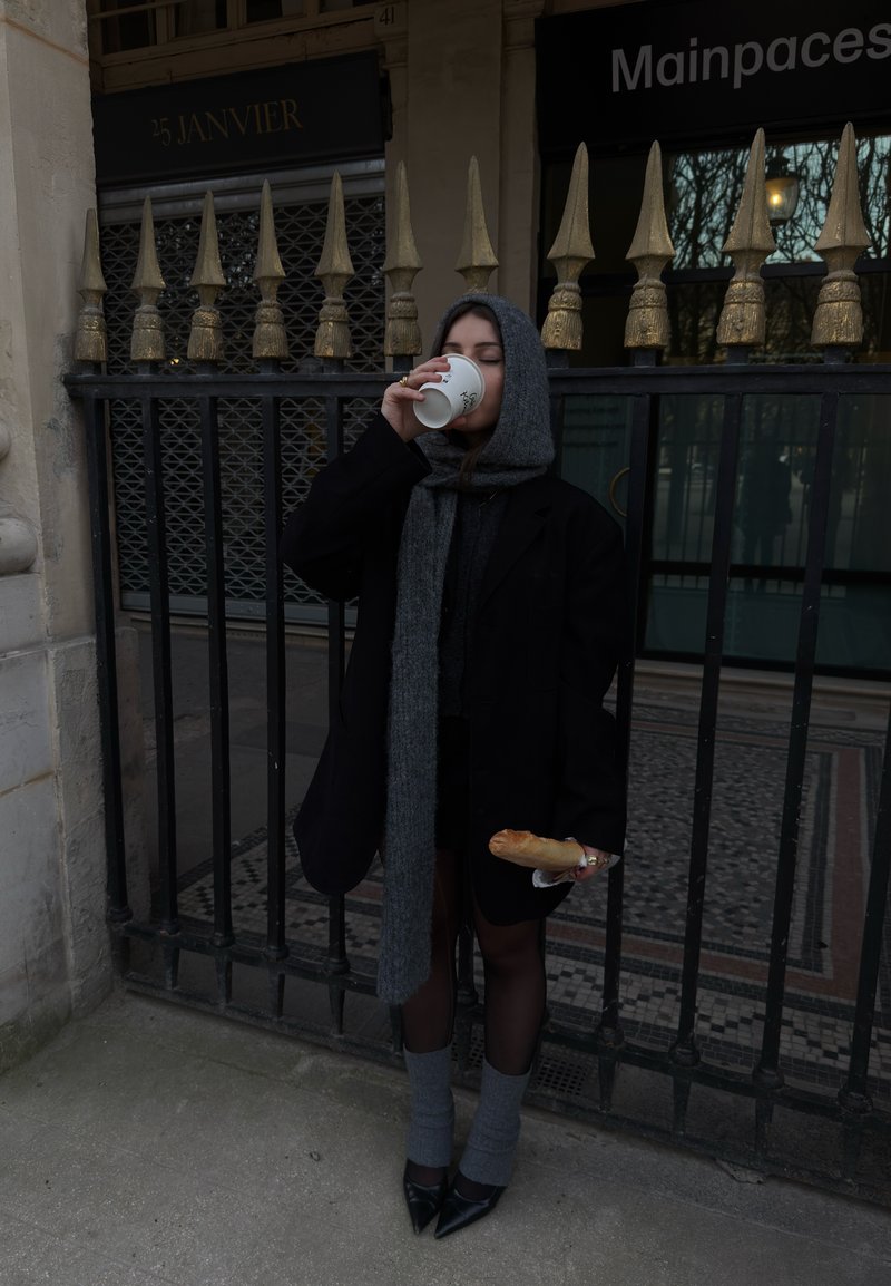 Kvinde iført grå hætte og tørklæde drikker af en kop, mens hun holder et baguette foran et sort jernhegn med gyldne spidser.