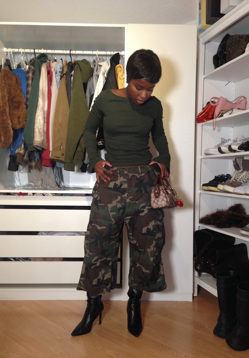 Persona con pantaloni mimetici, top verde scuro, stivali neri con tacco, che tiene una piccola borsa marrone all'interno di un armadio con vestiti appesi.