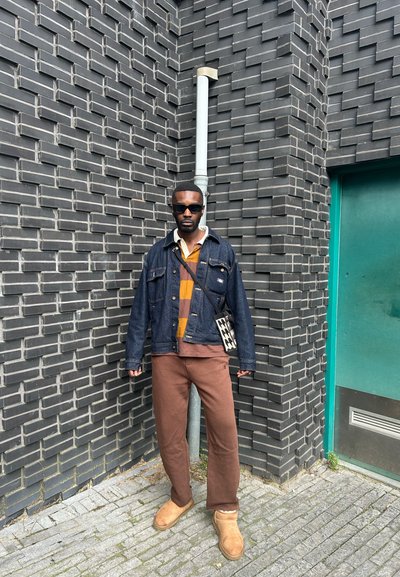 Chaqueta de mezclilla sobre una camisa a rayas marrón y mostaza, pantalones marrones y botas beige. De pie contra una pared de ladrillo oscuro texturizado y una puerta verde.