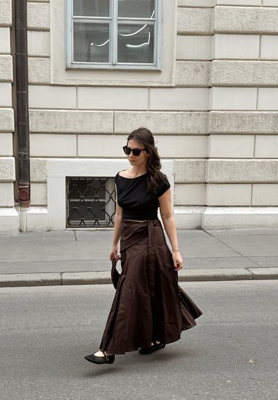 Mujer con top negro, falda larga marrón y gafas de sol caminando por una calle de la ciudad junto a un edificio de piedra beige.