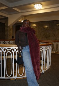 Femme aux longues tresses appuyée contre une rambarde blanche à l'intérieur, portant un haut marron, un jean bleu, des gants noirs, tenant un sac noir et une grande écharpe bordeaux.