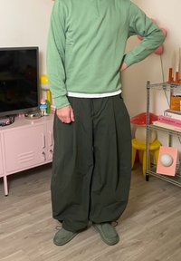 Pulover verde cu fermoar peste o cămașă albă, asortat cu pantaloni largi verde închis și pantofi gri. Fundalul prezintă decor colorat.