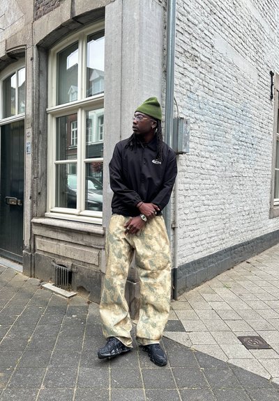 Chaqueta negra con logo, gorro verde, pantalones beiges de gran tamaño con un patrón mármol, y calzado negro con suelas texturizadas. Entorno urbano.