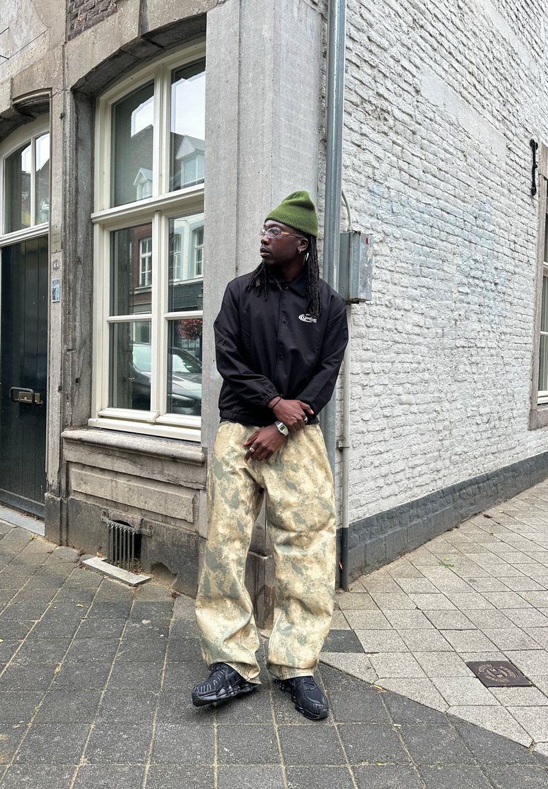 Svart jacka med logotyp, grön beanie, överdimensionerade beige byxor med marmorering, och svarta skor med strukturerade sulor. Urban miljö.