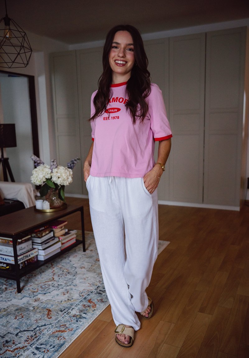 Jeune femme souriante, portant un t-shirt rose et un pantalon blanc, debout dans un salon avec un sol en bois et une table basse avec des livres et des fleurs.