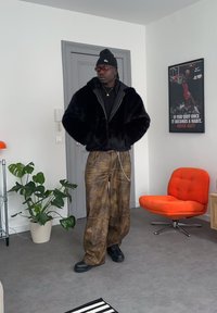 Homme portant un bonnet noir, des lunettes de soleil, une veste en fourrure noire, un pantalon ample marron avec chaîne, debout dans une pièce avec une chaise orange et une affiche de basket-ball.