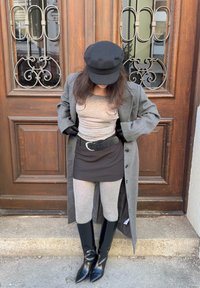 Manteau gris, chemise grise, jupe noire, leggings gris et bottines noires à bouts pointus. Casquette noire et gants complètent la tenue. Porte en bois en arrière-plan.