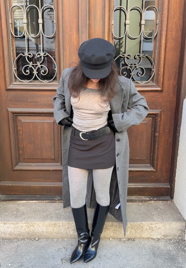 Szary płaszcz, szara koszula, czarna spódnica, szare legginsy i czarne szpiczaste buty. Dopełnieniem stroju jest czarna czapka i rękawiczki. Tło stanowi drewniane drzwi.