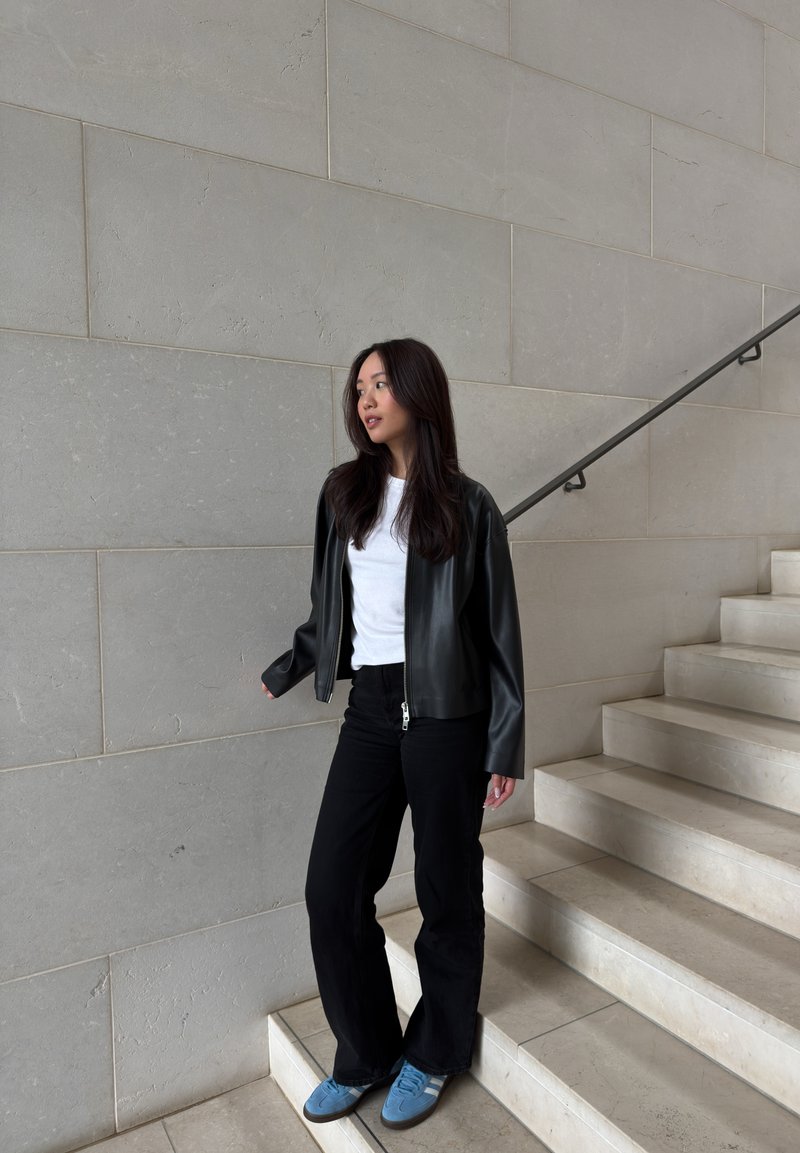 Jeune femme aux longs cheveux foncés se tenant sur des escaliers, portant une veste en cuir noire, une chemise blanche, un pantalon noir et des baskets bleues devant un mur en pierre.