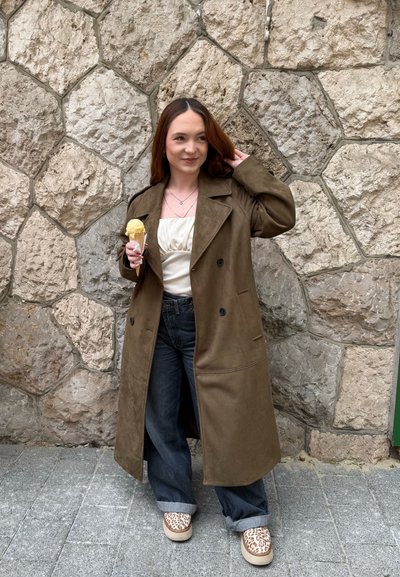 Joven mujer con un abrigo largo marrón, camiseta blanca y vaqueros, sosteniendo un cucurucho de helado, de pie junto a una pared de piedra.