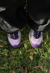 Lila és rózsaszín színű sportcipők fekete részletekkel, texturált hálós felsőrésszel és vastag fehér talppal, fentről nézve, fűre helyezve.