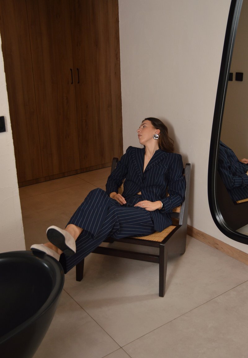 Femme en costume bleu marine à fines rayures, allongée sur une chaise en bois à côté d'un grand miroir et d'une armoire en bois dans une pièce minimaliste au sol carrelé.