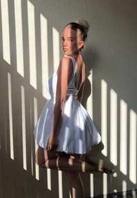 Robe blanche avec un corsage ajusté et une jupe évasée, présentant un dos plongeant. La lumière naturelle crée des motifs d'ombre rayés sur le mur.