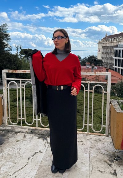 Suéter rojo de manga larga, cuello alto gris debajo, falda negra de cintura alta, cinturón negro con herrajes plateados, zapatos negros, entorno al aire libre.
