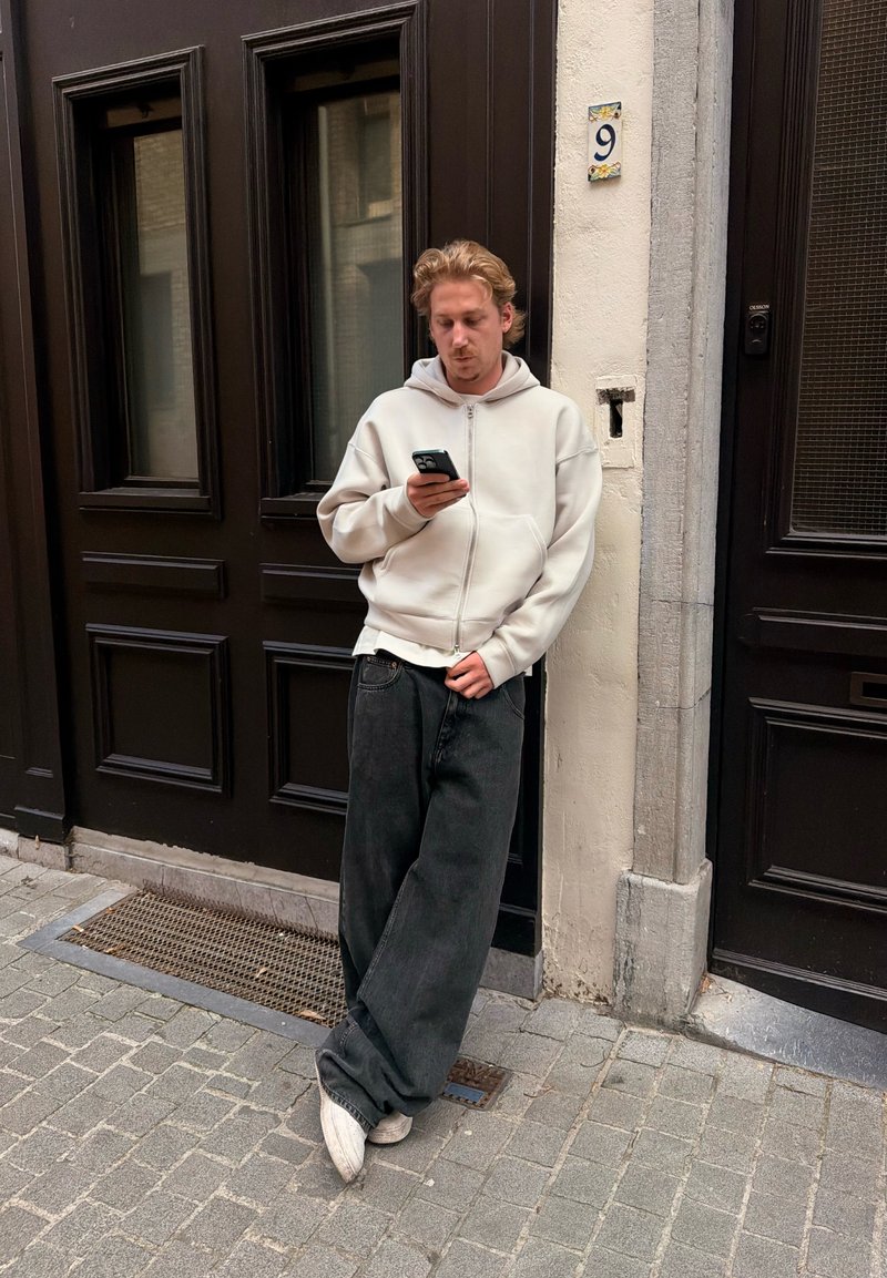 Jeune homme en sweat à capuche blanc et jean noir ample appuyé contre un mur de bâtiment, regardant son smartphone, près d'une porte avec le numéro 9 sur un mur clair.