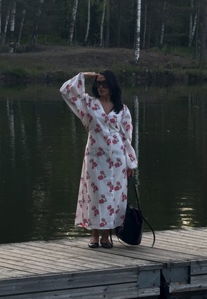 Largo vestido blanco con estampado floral rosa, mangas sueltas y escote en V. Bolso negro y zapatos planos. De pie en un muelle de madera junto al agua.