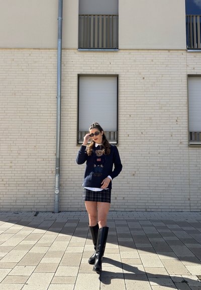 Mujer con gafas de sol, suéter azul marino con gráfico de oso, falda de cuadros y botas negras hasta la rodilla caminando por una calle pavimentada frente a un edificio de ladrillo claro.