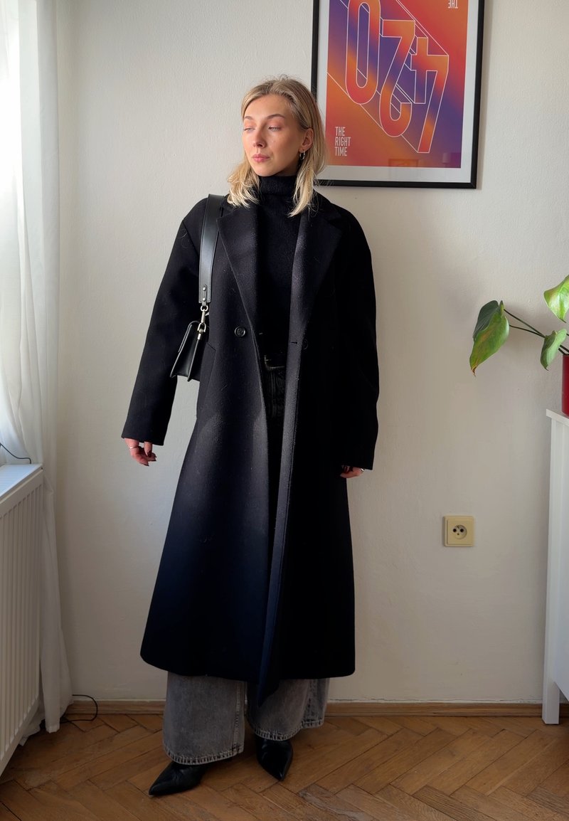 Jeune femme en long manteau noir, col roulé noir, jean gris à jambes larges et chaussures noires pointues, debout à l'intérieur près d'une fenêtre et d'une affiche encadrée.
