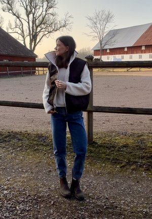 Mujer con chaleco negro, suéter crema, jeans y botas, de pie junto a una cerca de madera con graneros y árboles sin hojas al atardecer.