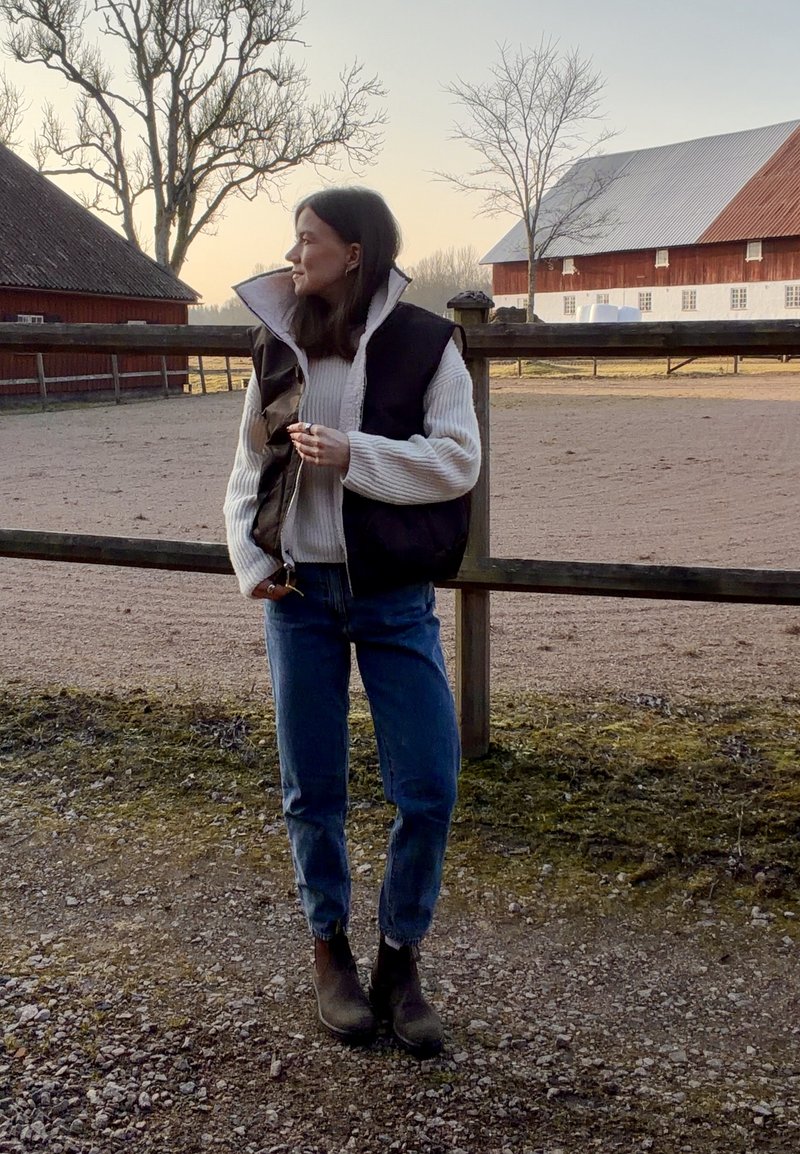 Kvinde iført sort vest, cremefarvet sweater, jeans og støvler står ved et træhegn med ladebygninger og nøgne træer ved solnedgang.