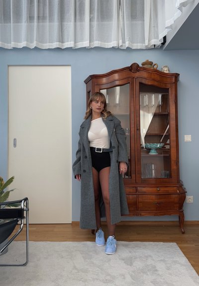 Abrigo largo gris de lana sobre una blusa blanca y pantalones cortos negros. Zapatillas azul claro, armario de madera al fondo, cortinas blancas y pared beige.