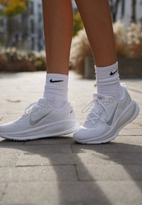 Ténis atléticos brancos com parte superior em malha respirável, detalhes prateados e sola almofadada. Usados com meias brancas caneladas com um logotipo preto.