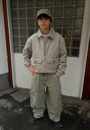 Persona con gorra beige, chaqueta beige con bolsillos delanteros, pantalones cargo amplios y botas negras, de pie junto a una pared de bloques de vidrio.