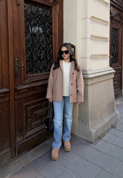 Abrigo beige de corte amplio, suéter crema, jeans azul claro y botas plataforma color tostado. Unas gafas de sol negras y un pequeño bolso negro completan el conjunto.